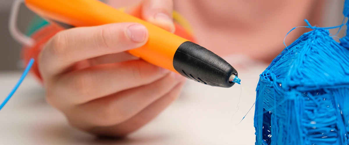 A hand holds an orange 3D printing pen while fine strands of blue filament emerge from the tip. In the foreground, a model made of blue plastic can be seen being designed with the pen. The focus is on the precise handling of the pen and the details of the model.