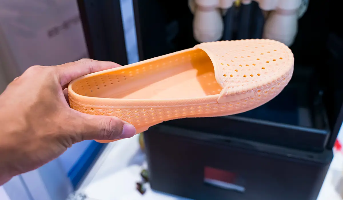 A hand holds an orange shoe made with a 3D printer. The shoe features a perforated surface and precise details that demonstrate the possibilities of additive manufacturing. In the background, a 3D printer can be seen out of focus, highlighting the production process.