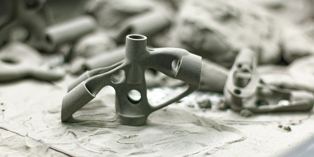 Various 3D printed metal parts lying in grey dust. The parts have complex geometric structures and were apparently manufactured using metal 3D printing technology.