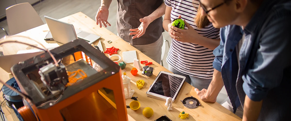 A group of people are standing around a table where a 3D printer is working. Various 3D printed objects, such as geometric shapes and figures, are lying on the table. The people seem to be discussing the printing process, while laptops and tablets can also be seen on the table.