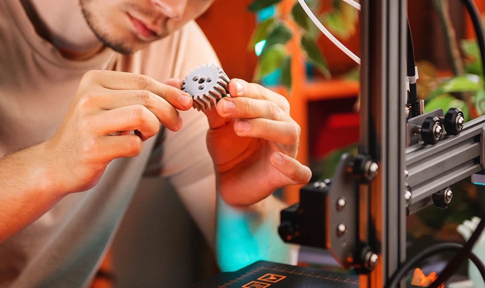 A man holds and examines a 3D printed gear. The printer that produced the gear is visible in the background.