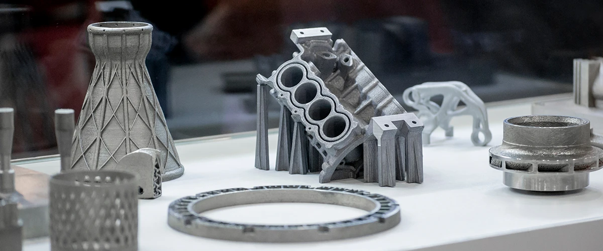 A collection of 3D printed metal parts, including engineering components such as an engine block and a lattice structural element. The metal parts demonstrate the precision and versatility of modern 3D printing technology.