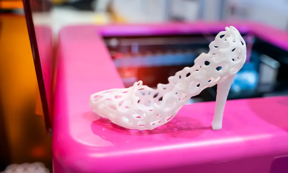 Ein weißer High Heel mit einer filigranen, wabenartigen Struktur steht auf einem pinkfarbenen Untergrund. Der Schuh wurde mit einem 3D-Drucker hergestellt und zeigt die präzisen und kreativen Möglichkeiten dieser Technologie. Im Hintergrund ist unscharf der 3D-Drucker zu erkennen, der an der Fertigung beteiligt war.