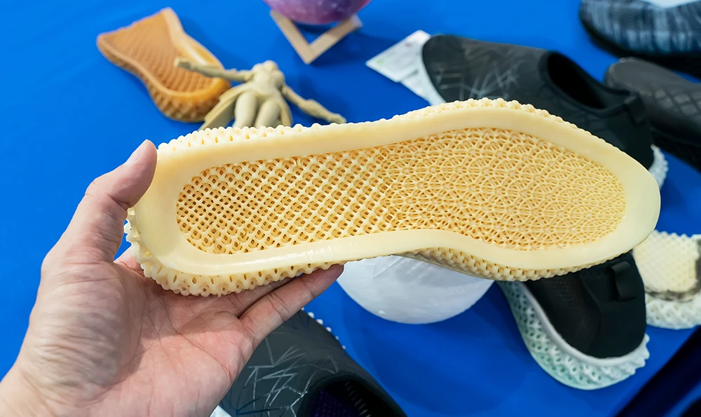 A person holds a 3D printed shoe sole with a honeycomb structure in his hand, while various shoes and soles lie on a table in the background.