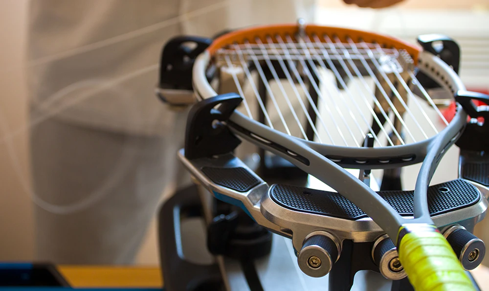 A tennis racket is restrung on a stringing machine while a person works on it.