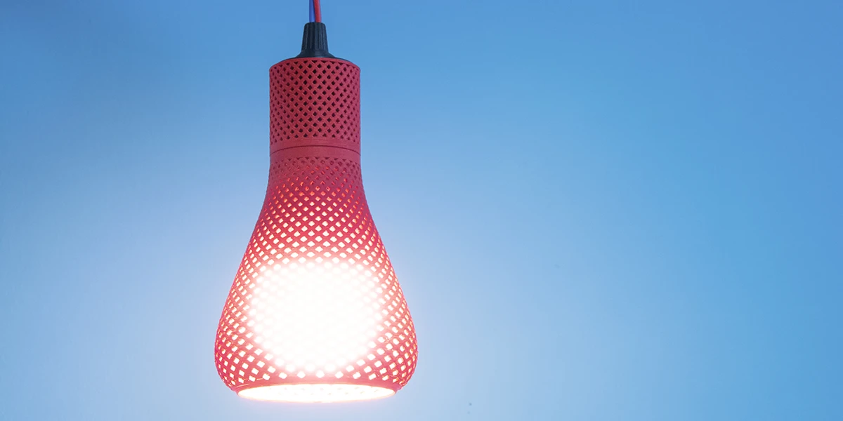 A 3D printed lamp in the shape of a drop made of a red perforated material. It is attached to a red cable suspension and emits warm light.