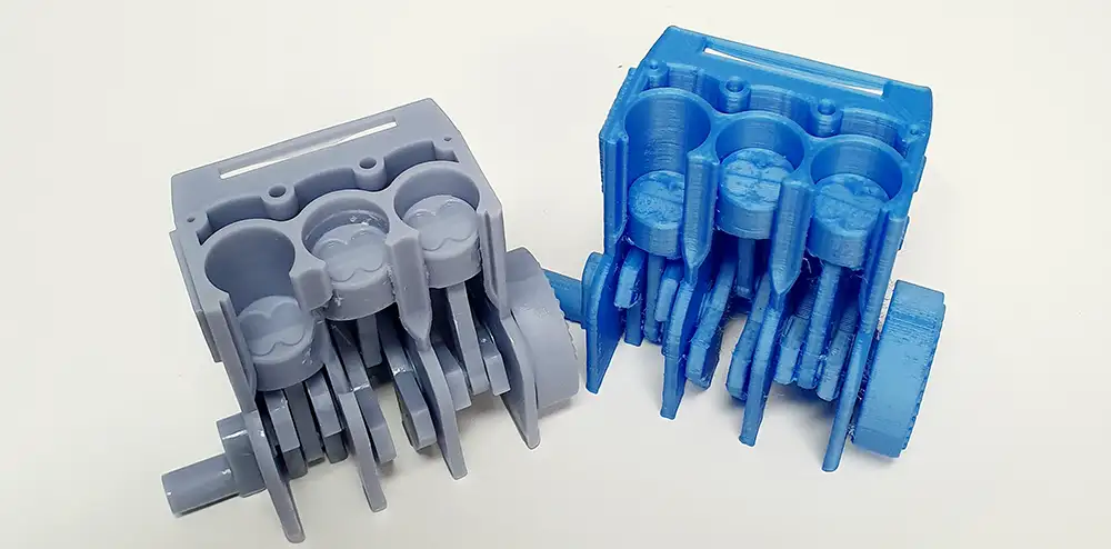 Various 3D printed models of an engine block, a grey part and a blue part made of plastic material.