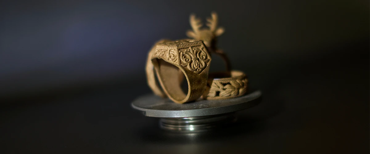 The image shows detailed gold-colored jewelry, presumably rings, displayed on a platform. The rings have elaborate engravings and a complex design, possibly resulting from a 3D printing process or traditional jewelry craftsmanship.