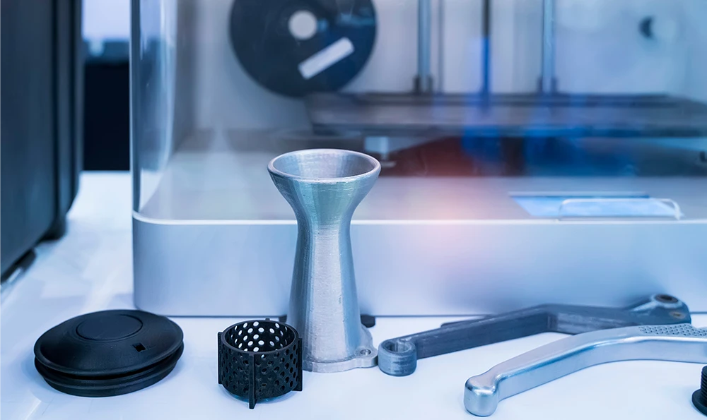 Various 3D printed parts lie on a table next to a 3D printer. Among them are a funnel-shaped object, a perforated grid, a tool and other metal parts.