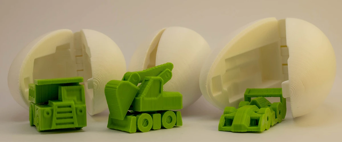 Three white 3D printed eggs lie open on a surface, and inside each egg is a small green 3D printed vehicle. The vehicles are richly detailed, including a truck, a crane and a race car. This set shows the creativity and precision that is possible with 3D printing.