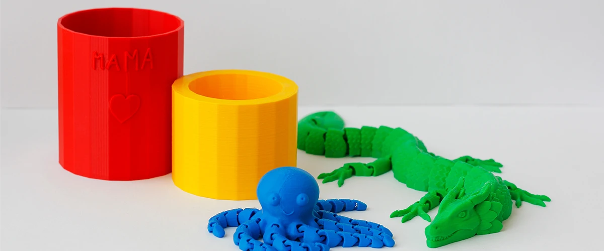 On a white surface, several colorful 3D printed objects stand: a red mug engraved with "Mama" and a heart, a yellow mug, a blue, smiling octopus and a green dragon with movable limbs. The detailed design and bright colors show the versatility of 3D printing.