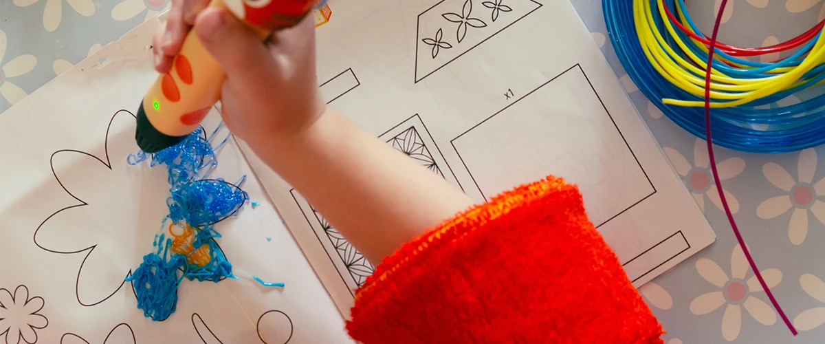 A child works with a 3D printing pen and creates a template with colorful plastic. The hand holds the pen while blue and yellow structures are drawn on the template. On the right you can see colorful filaments that are used for the 3D printing pen.