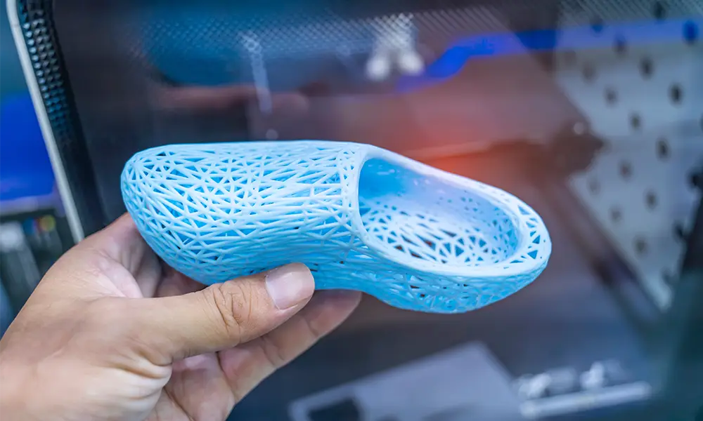 Eine Hand hält einen hellblauen Schuh mit einer offenen, netzartigen Struktur, der mit einem 3D-Drucker hergestellt wurde. Der Schuh ist leicht und innovativ gestaltet und zeigt die Möglichkeiten moderner 3D-Druck-Technologie. Im Hintergrund ist unscharf der 3D-Drucker sichtbar, der das Modell produziert hat.