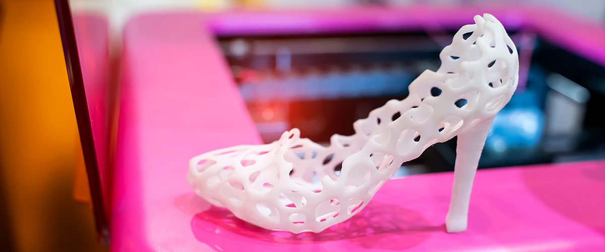 A white high-heeled shoe with a complex, net-like design stands on a pink surface. The shoe was made using a 3D printer and has a futuristic and avant-garde aesthetic.