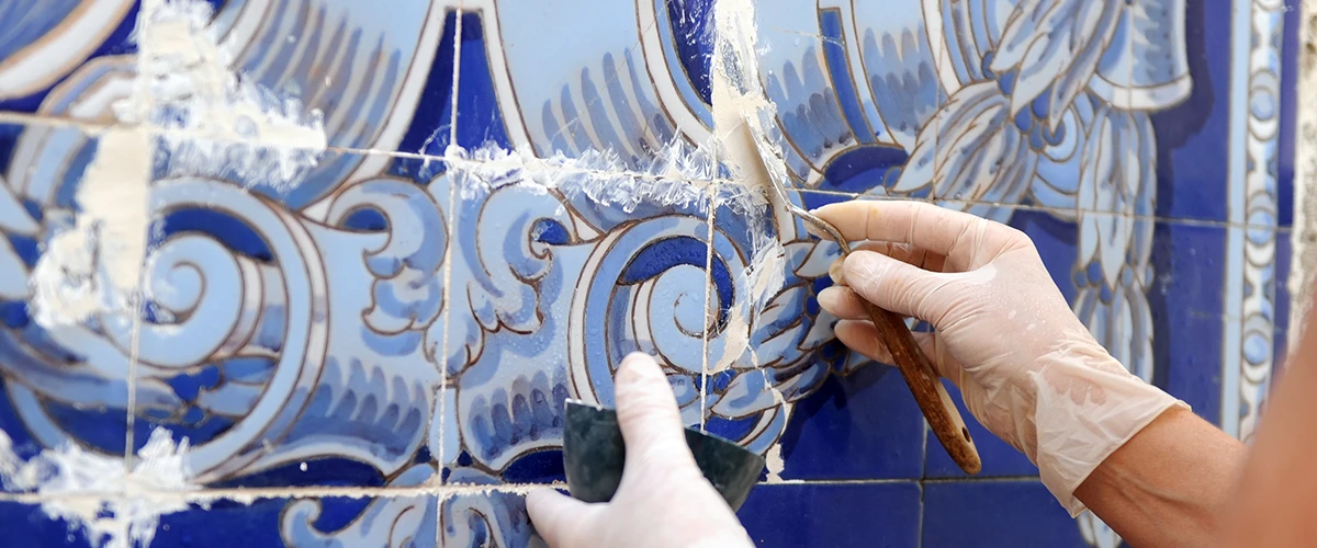 Two hands wearing gloves work on restoring a blue tile wall with intricate patterns. They use tools to work on the grout and repair damaged areas.