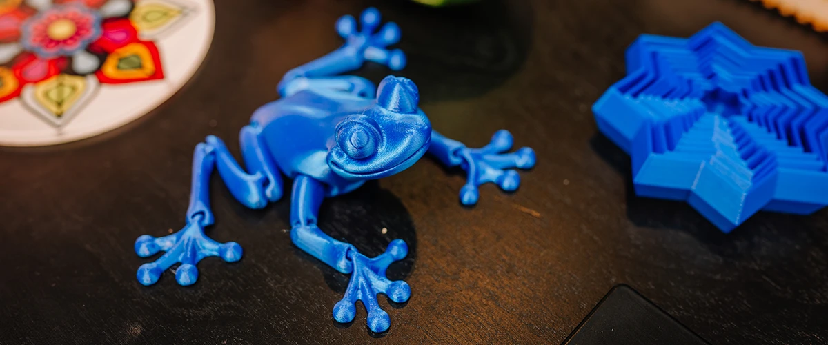 A blue 3D printed frog figure lies on a dark surface, impressing with its detailed design and realistic shape. Next to it are other colorful 3D printed objects, including a geometric structure and a decorative design. The scene shows the diversity and creativity in 3D printing.