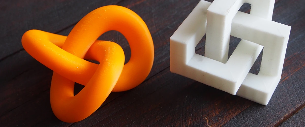 Zwei 3D-gedruckte Objekte liegen auf einer dunklen Holzoberfläche: ein orangefarbenes, verschlungenes Knotenmuster und eine weiße, geometrische Struktur mit ineinander verschachtelten Quadraten. Beide Modelle zeigen die Präzision und Vielseitigkeit des 3D-Drucks bei der Erstellung von komplexen Formen.