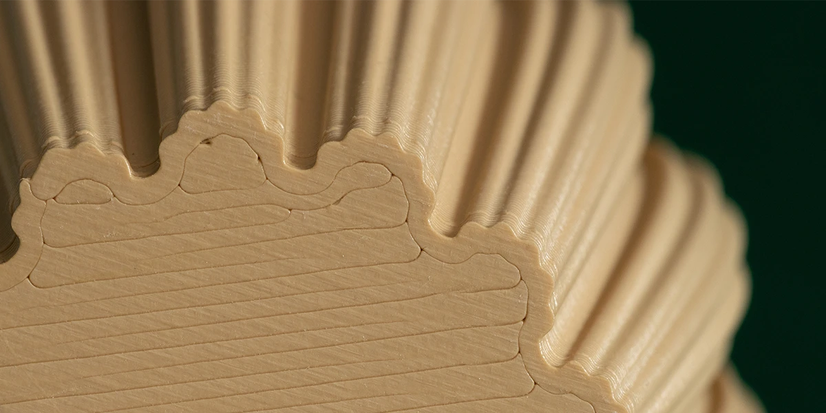 Close-up of a 3D printed object showing a textured surface with layers of thin lines created by the printing process. The material is beige in color and the surface is arranged in wavy lines.