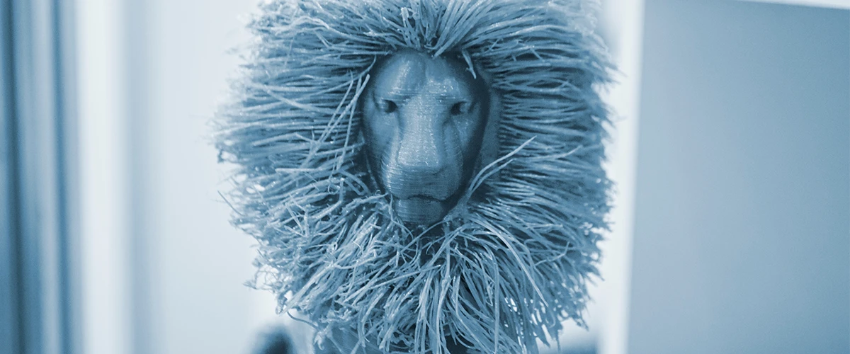 A 3D printed model of a lion's head with a detailed, wispy mane is shown. The mane consists of fine, fibrous structures that give the impression of realistic hair.