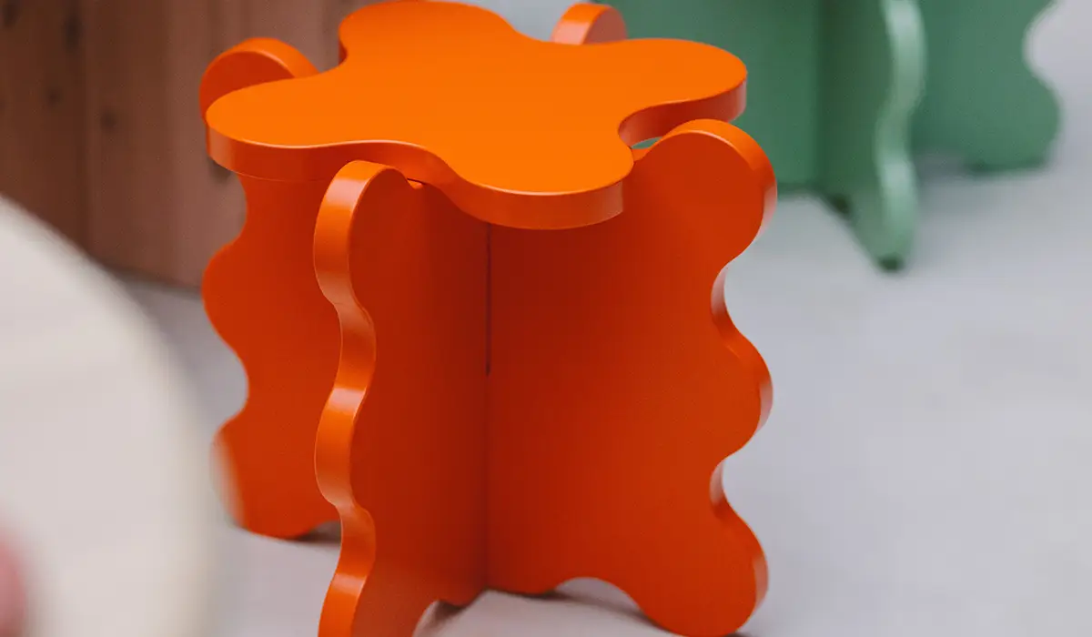 A 3D printed chair with a striking, organic shape in bright orange. The stool has a wavy structure on the sides, giving it a modern and playful look.