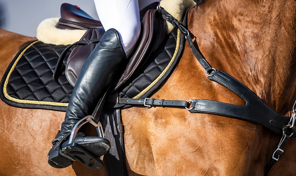 A rider in black boots sits in the stirrup on a saddled brown horse with a black saddle blanket and bridle.