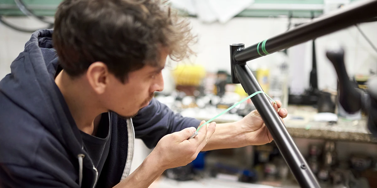 A man is working on a bicycle frame and marking a spot with a green tape. The background shows a workshop environment equipped with various bicycle parts and tools.