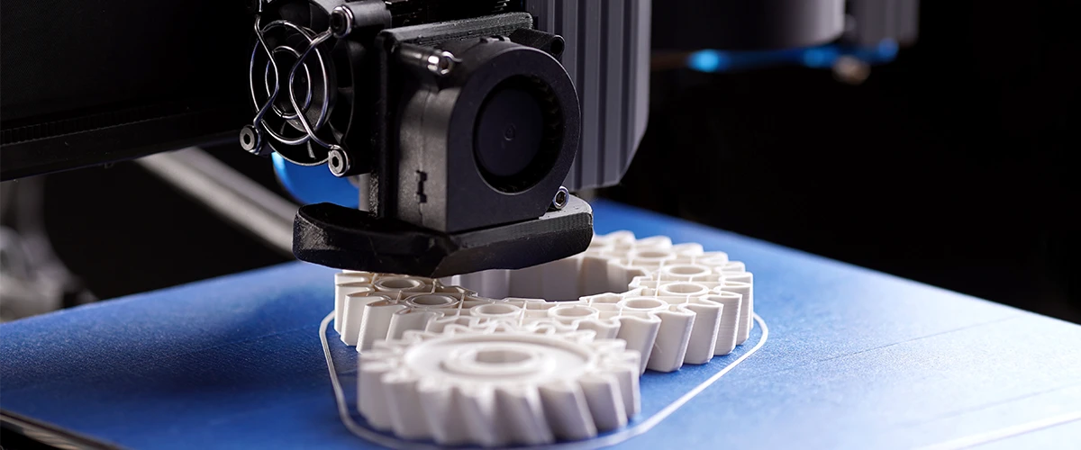 Close-up of a 3D printer creating a complex white gear model. The print head moves precisely over the blue build plate as the workpiece is built layer by layer.