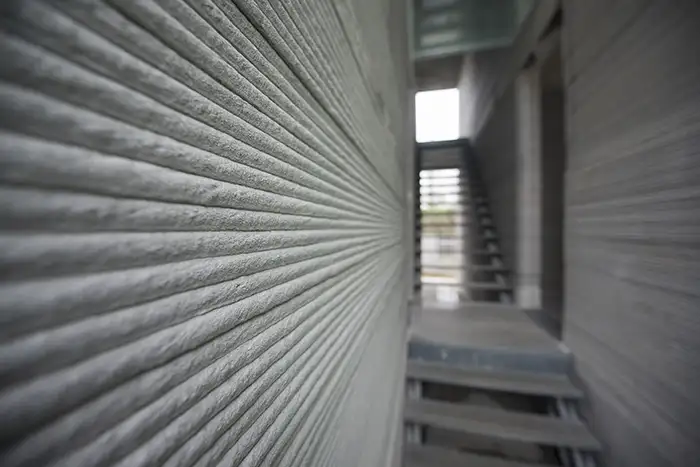 Close-up of a wall of a 3D-printed concrete building, showing the textured layers. A staircase leading to an upper floor can be seen in the background.