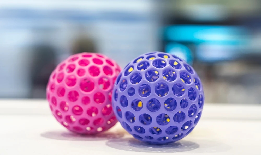 Two 3D-printed hollow balls with honeycomb-shaped openings in the colors pink and purple lie on a white surface.