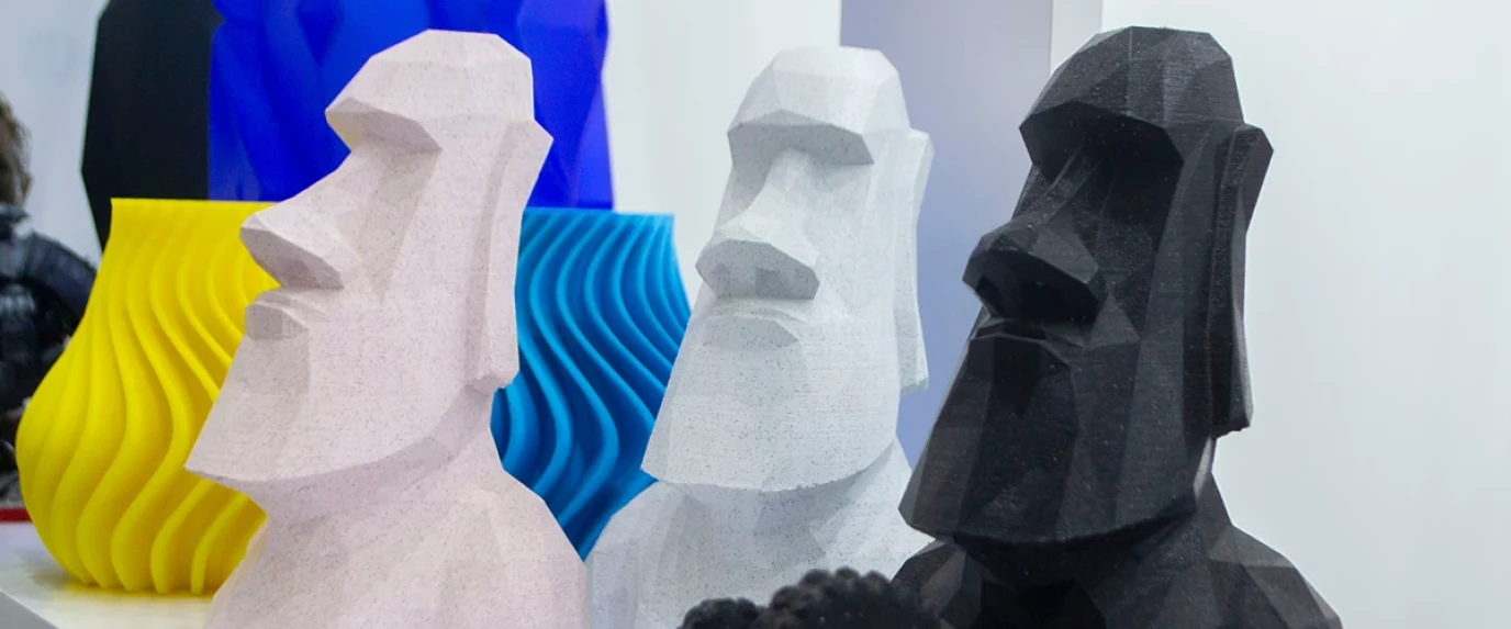 Mehrere 3D-gedruckte Statuen mit einem kantigen, abstrakten Design, das an die Moai-Statuen von Osterinsel erinnert, sind in verschiedenen Farben wie Schwarz, Weiß und Grau ausgestellt. Im Hintergrund sind weitere, wellenartige Skulpturen in Blau und Gelb zu sehen.