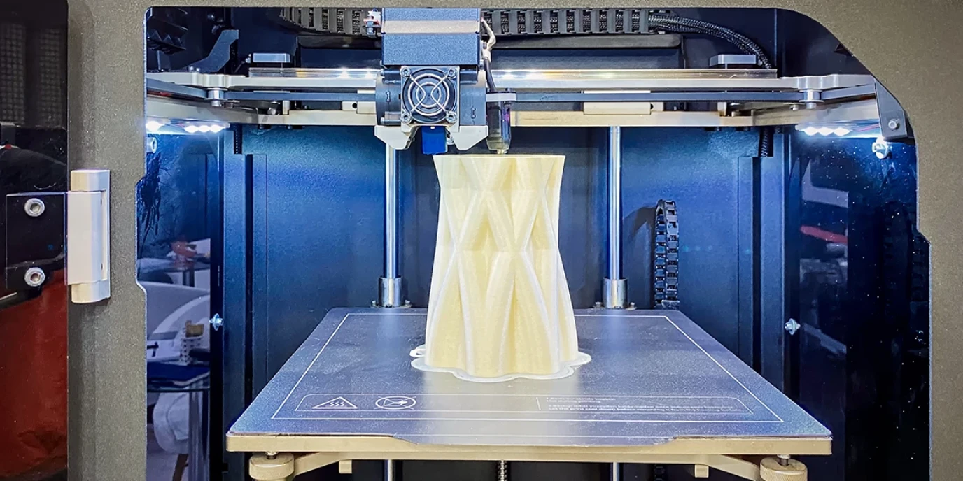 A 3D printer printing an object with a textured, geometric surface made of a light-colored material. The object is almost finished and is on the print bed.