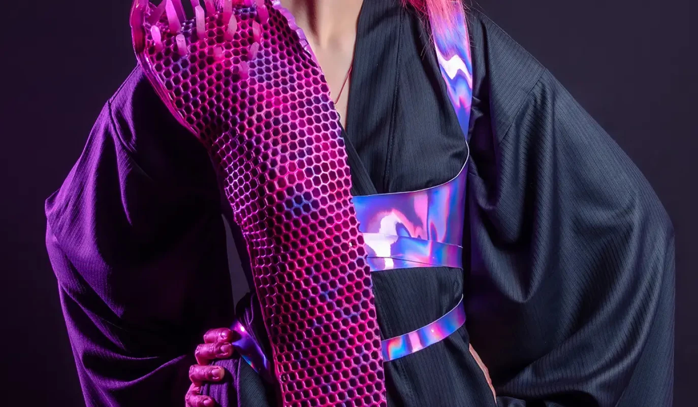 Eine Person mit leuchtend pinkem Haar trägt einen futuristisch anmutenden schwarzen Kimono, kombiniert mit einem auffälligen, netzartigen, pink-violetten Armstück, das durch 3D-Druck gefertigt wurde. Die Gesamtästhetik ist modern und erinnert an Cyberpunk-Mode.