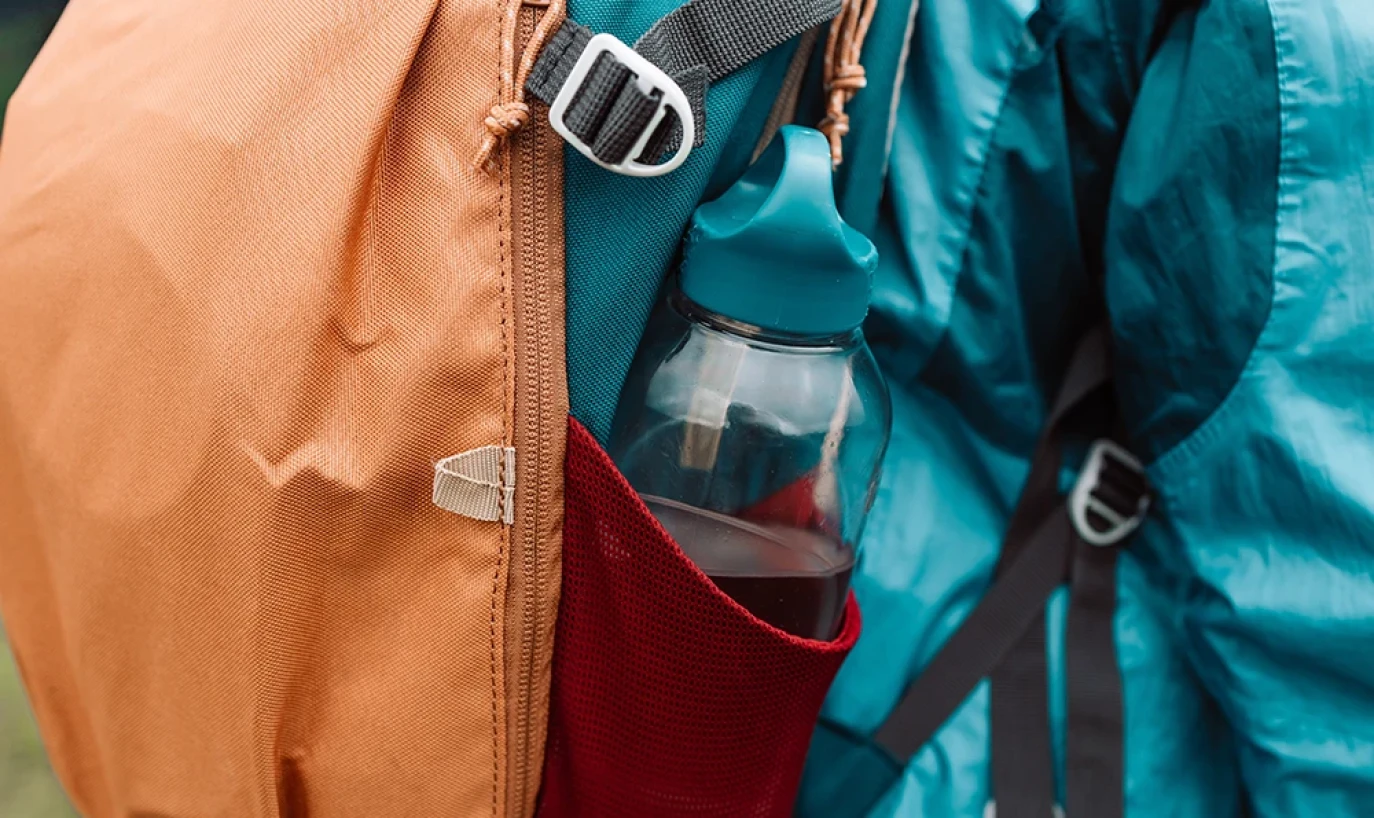 Ein Rucksack mit einer Wasserflasche in der Seitentasche wird von einer Person mit blauer Outdoor-Jacke getragen.