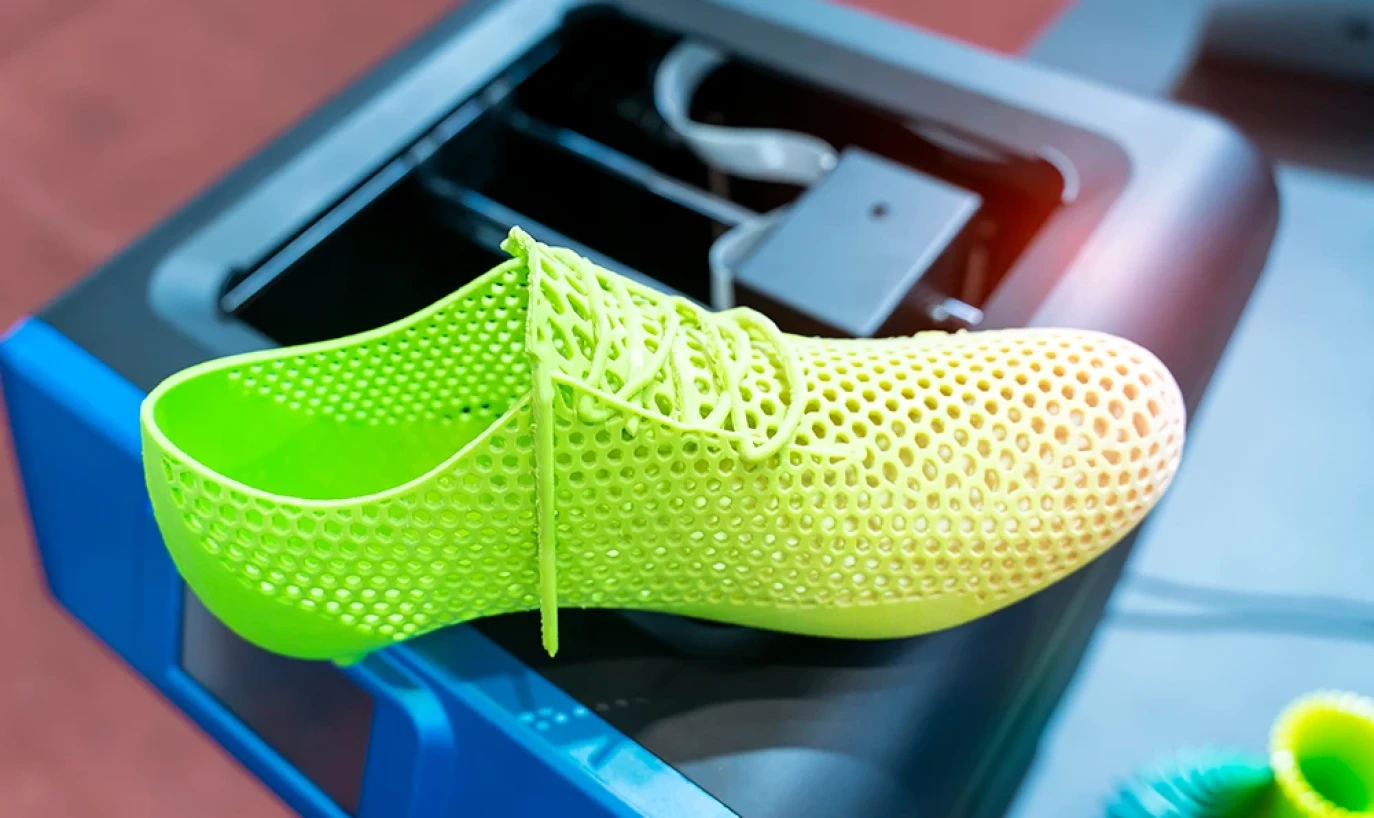 The image shows a 3D printed shoe with a honeycomb structure in bright color lying on a machine or printer.