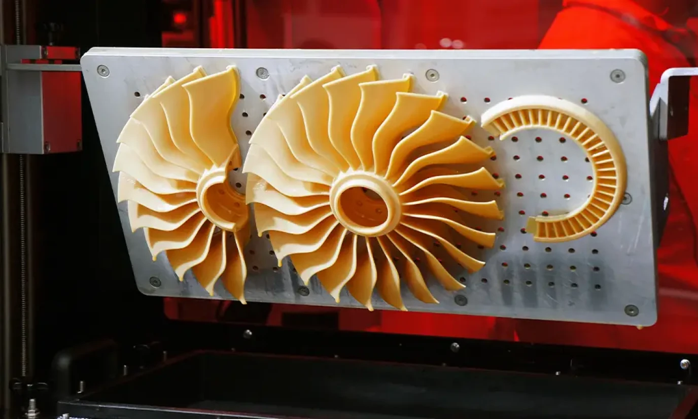 3D-printed prototypes of turbine blades in an industrial printer, showing aerodynamic designs made of plastic. The yellow color of the blades contrasts with the red illuminated environment of the printer.